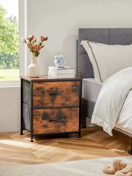 A brown nightstand with two fabric drawers, placed next to a bed in a bedroom setting, featuring a wooden tabletop with a clock and a vase of flowers.