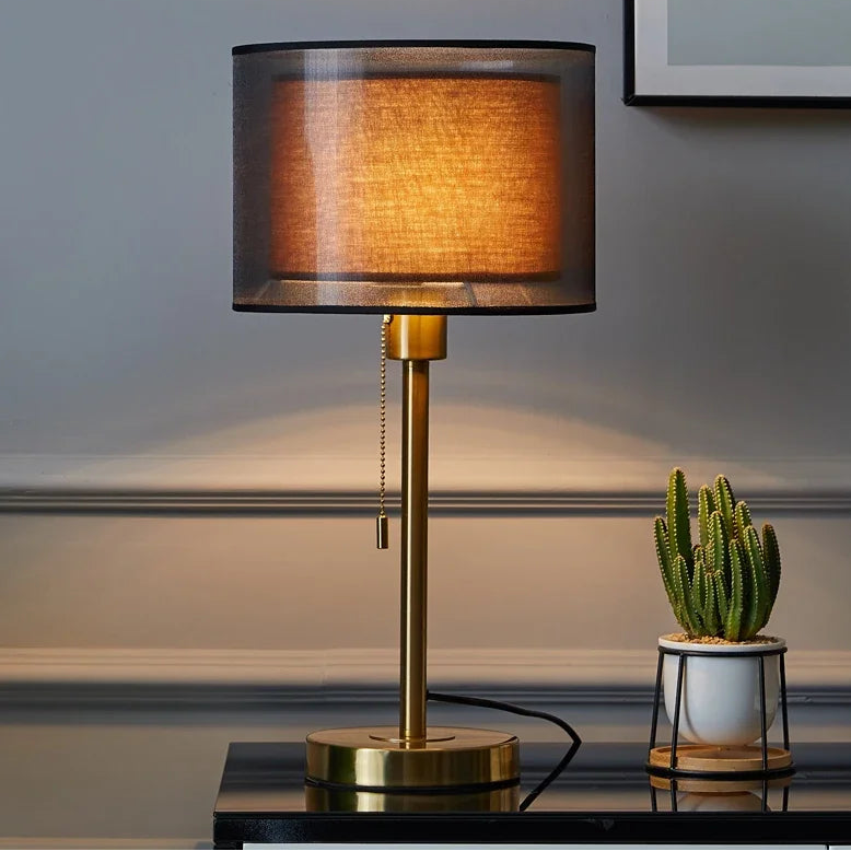 A gold-colored, modern style table lamp with a shade, placed on a flat surface with a small potted plant to the right.